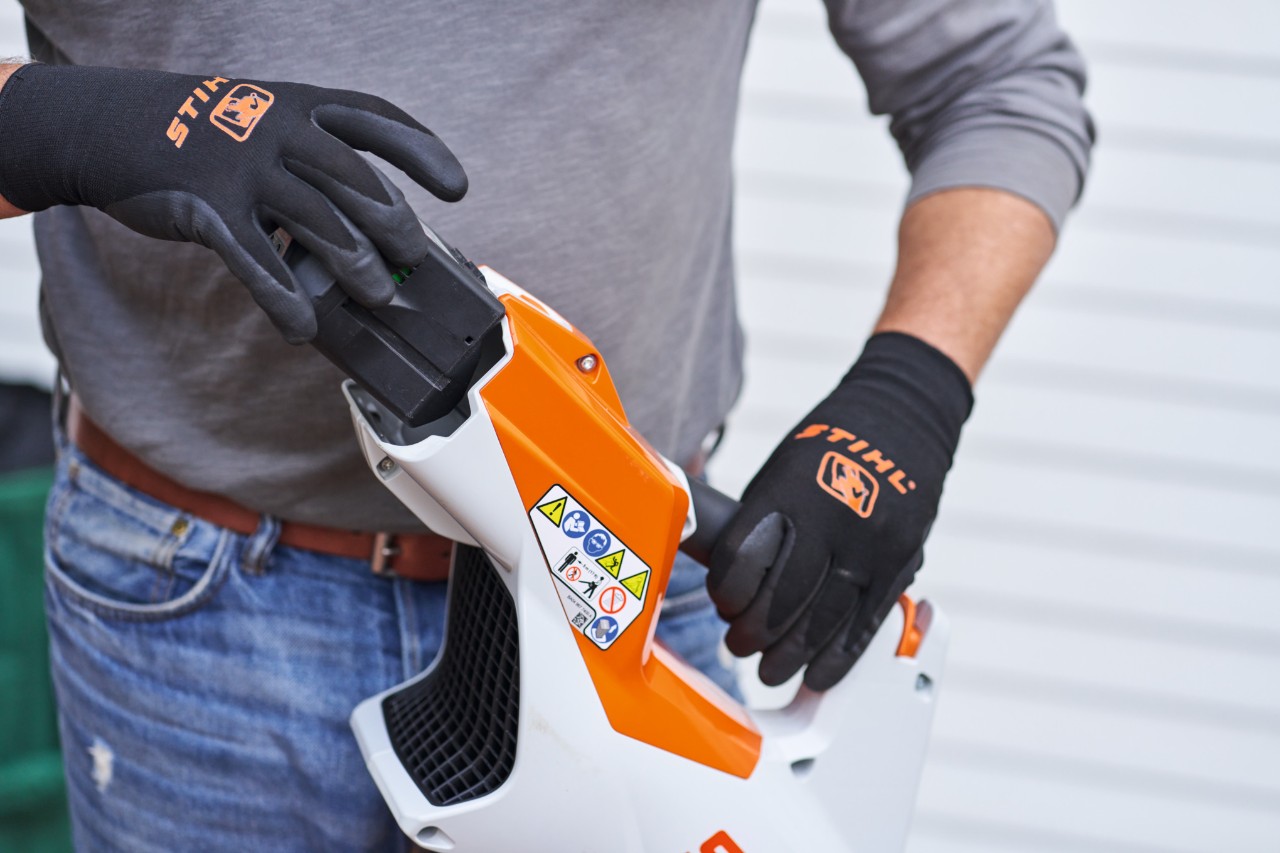 Egy fekete STIHL kesztyűt viselő ember kiveszi az akkumulátort a lombfúvóból