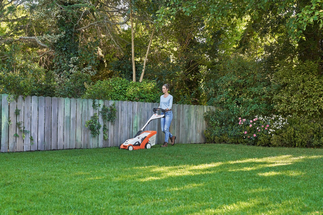 Egy nő STIHL RMA 339 C akkumulátoros fűnyíróval nyírja a gyepet egy fakerítés előtt.