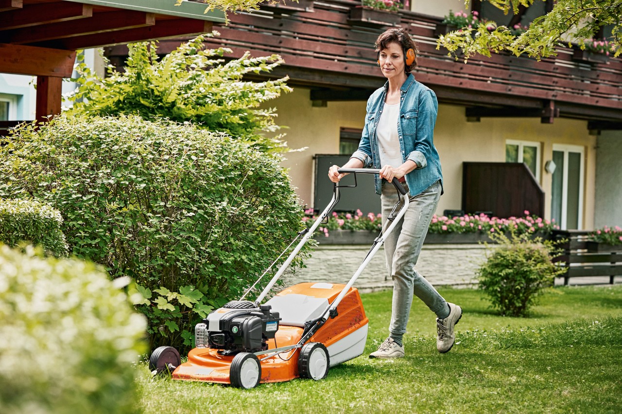 Egy nő tavasszal STIHL RM 243 T benzines fűnyíróval ápolj a a gyepet a ház előtt a kertben