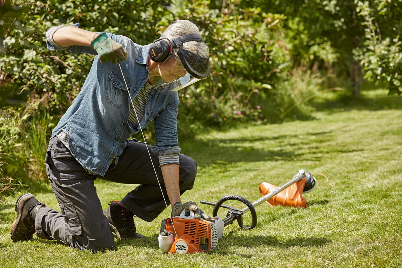Hallás- és arcvédőt viselő nő egy zöld területen STIHL FS 56 R benzines motoros kaszát készül elindítani