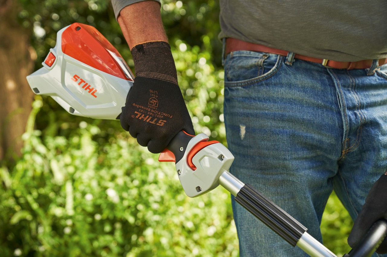 Védőkesztyűt viselő férfi megnyomja a STIHL FSA 60 akkumulátoros kasza kapcsolóemeltyű-zárát 