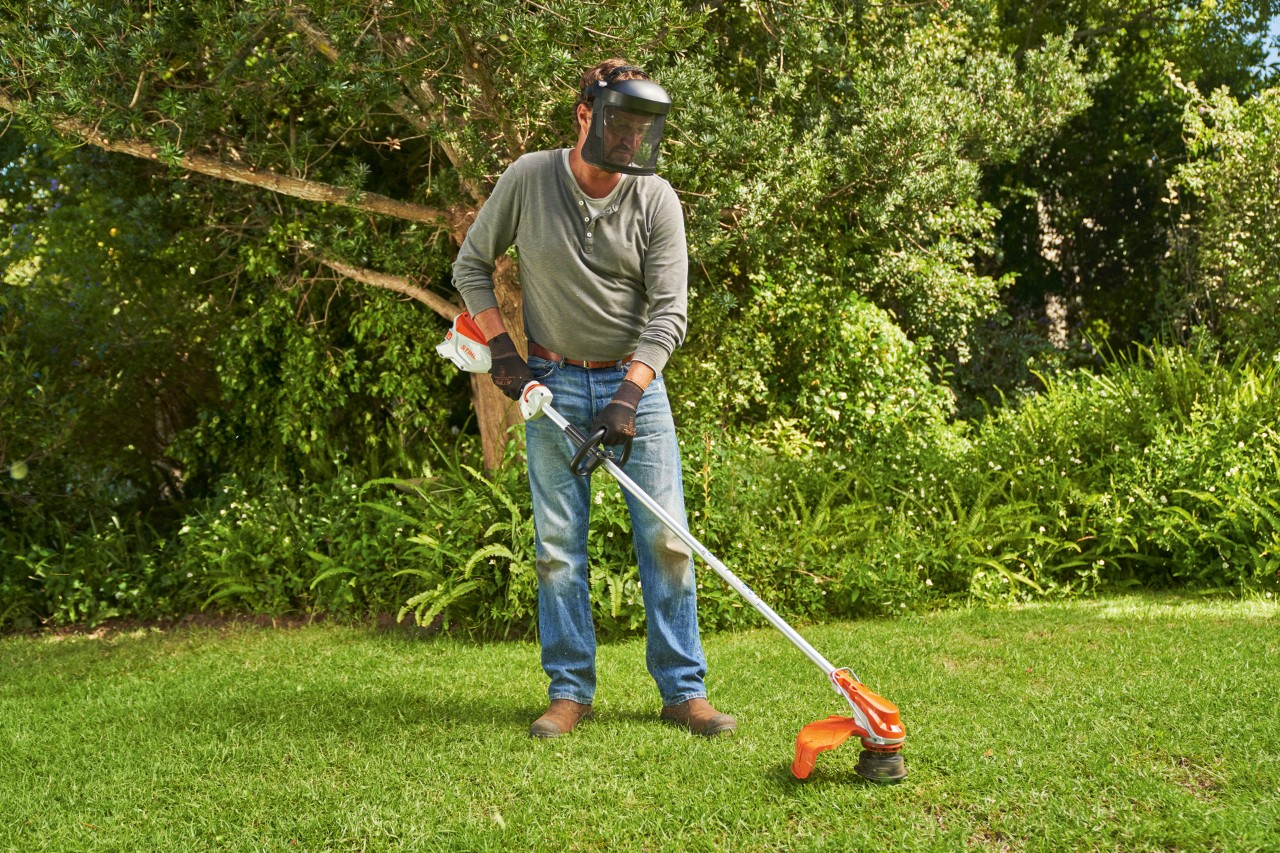 Egy arcvédőt viselő férfi STIHL FSA 60 akkumulátoros kaszával füvet nyír a kertben.