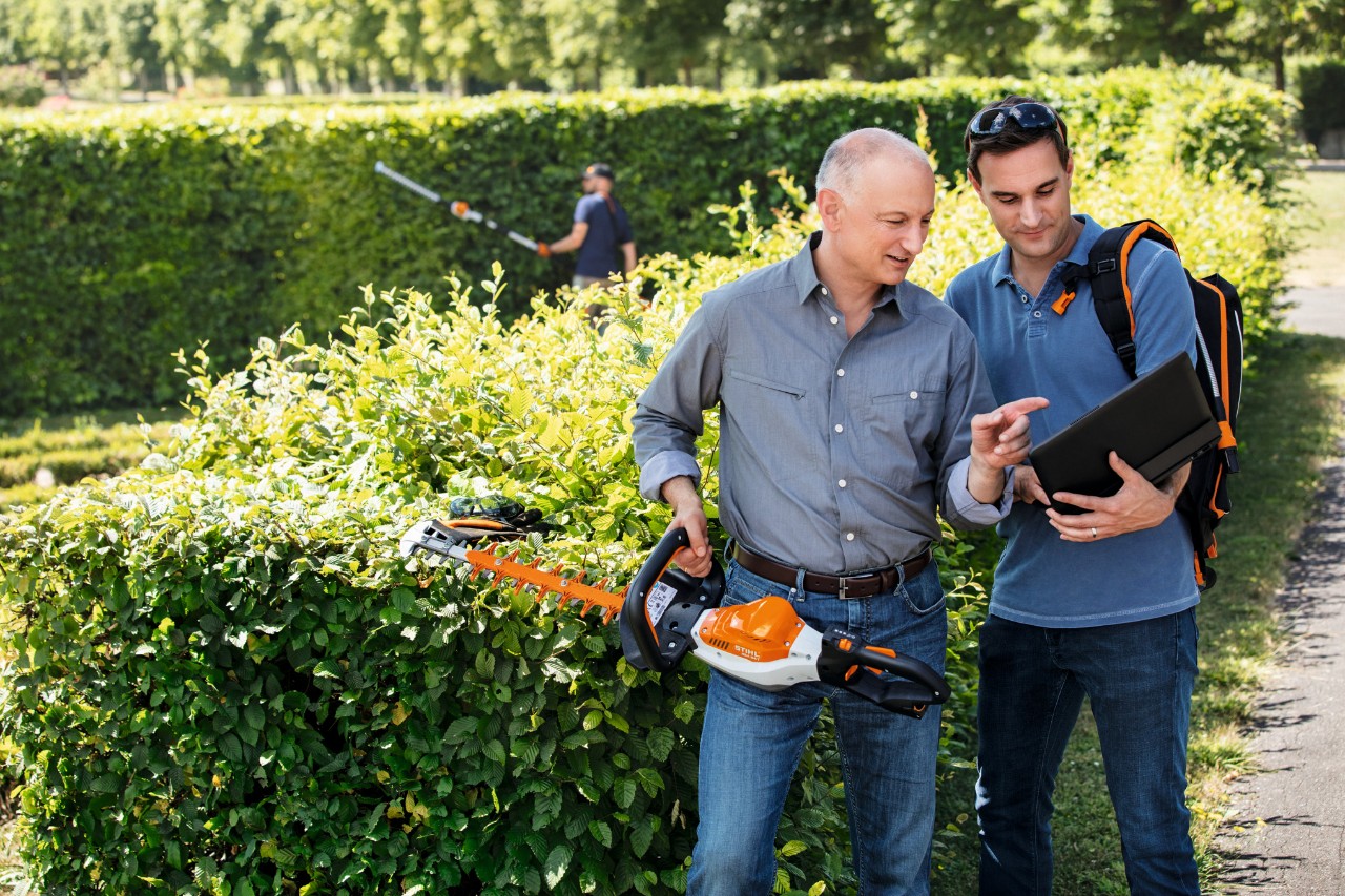 Dr. Nikolas Stihl holds a STIHL hedge trimmer in his hand and talks to a person standing next to him holding a laptop.
