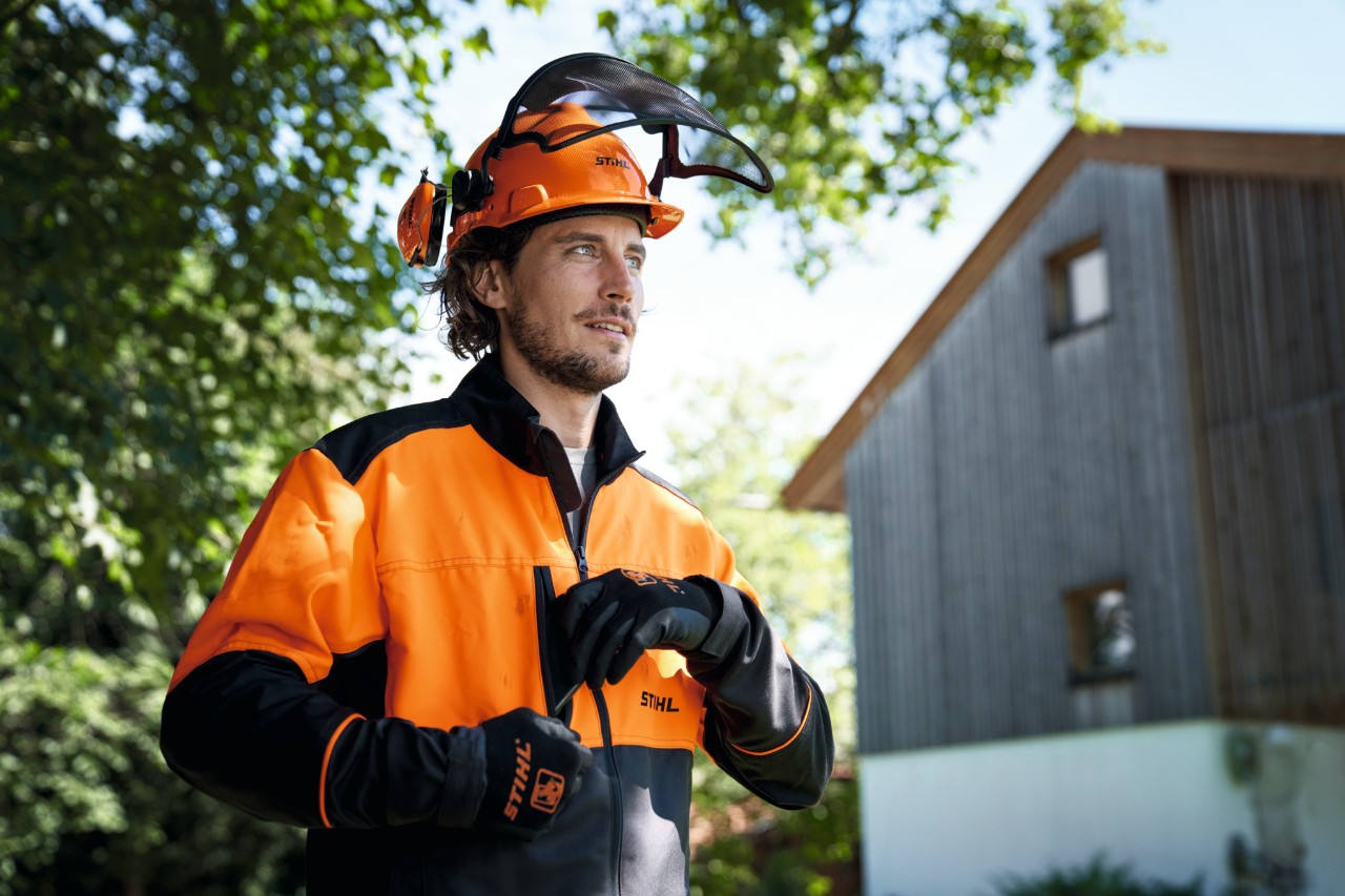 Férfi narancssárga és fekete STIHL védőeszközben, sisakkal és kesztyűvel, magabiztosan néz ki a képből, a háttérben egy ház és fák 