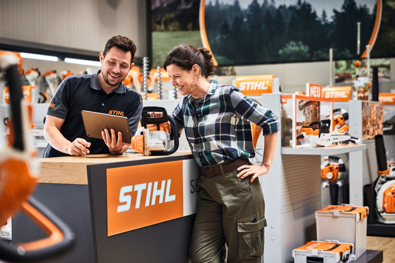 STIHL szakkereskedő STIHL sövénynyírót vásároló fiatal nőnek ad tanácsot a bemutatópolc előtt