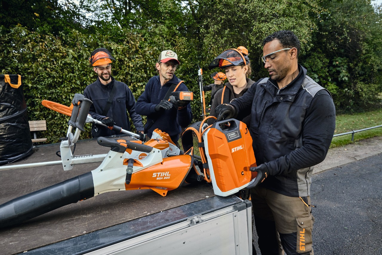 Három férfi és egy nő STIHL védőfelszerelésben egy jármű rakfelülete előtt áll, amelyen STIHL készülékek láthatók, többek között egy STIHL BGA 200 akkumulátoros lombfúvó és egy háton hordozható STIHL AR 2000 L akkumulátor 