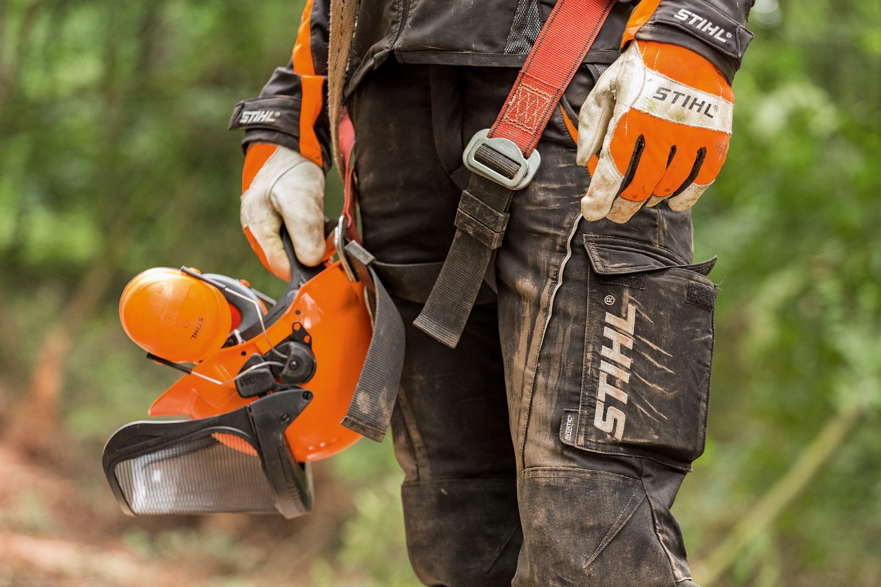 STIHL vágásbiztos nadrágot és STIHL kesztyűt viselő férfi STIHL védősisakot tart a kezében 