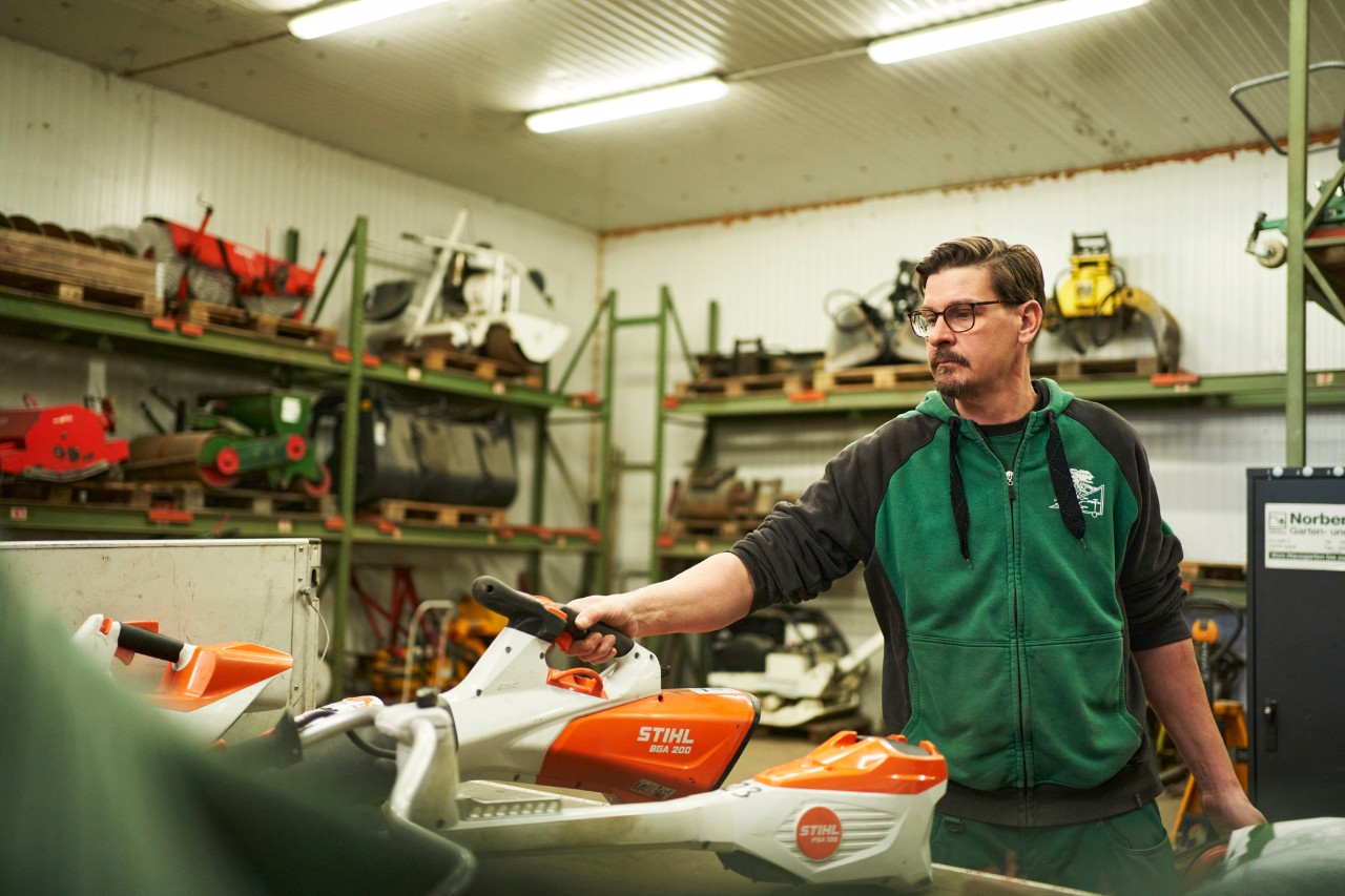 Egy férfi a műhelyben egy munkaasztal mögött áll, melyen STIHL akkumulátoros készülékek láthatók, kezében egy STIHL BGA 200 akkumulátoros lombfúvót tart