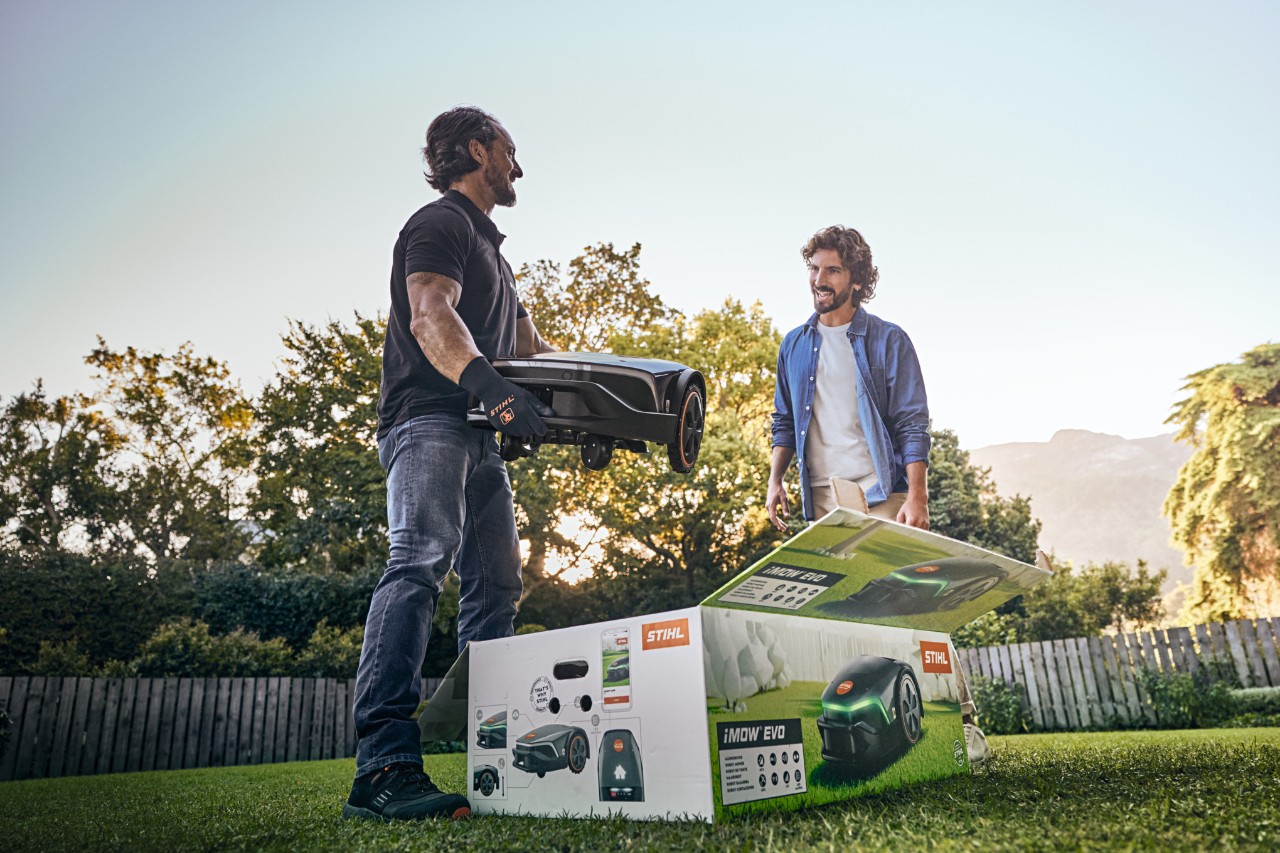 Egy STIHL szakkereskedő egy ügyfél kertjében áll, kezében egy iMOW® EVO-val, előtte az iMOW® dobozával, és segít az üzembe helyezésben