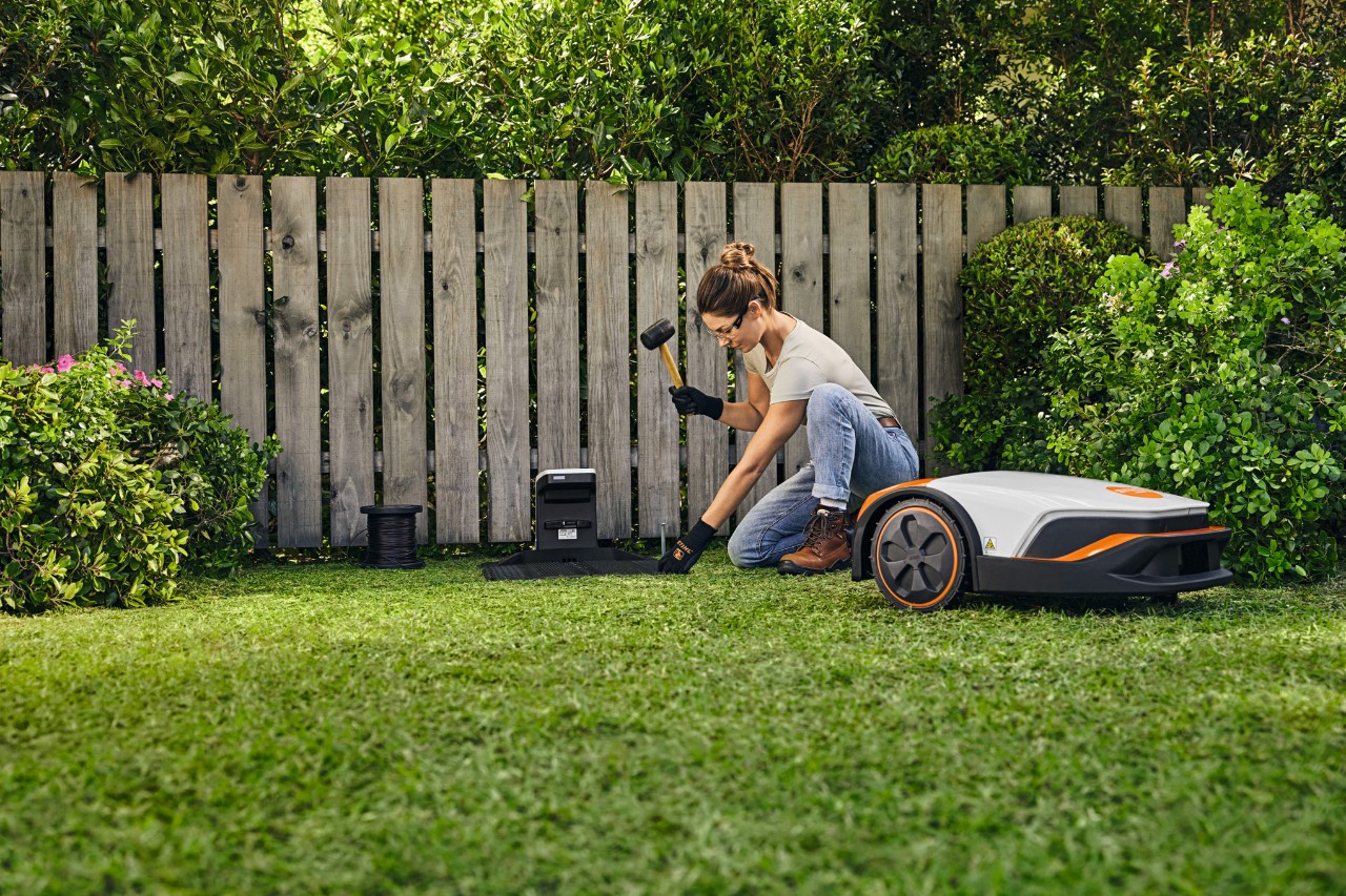 Egy nő egy STIHL iMOW® dokkolóállomást telepít a kertjében a kerítés előtt.
